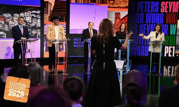 Five people stand at podiums in front of brightly coloured screens in a debate format.