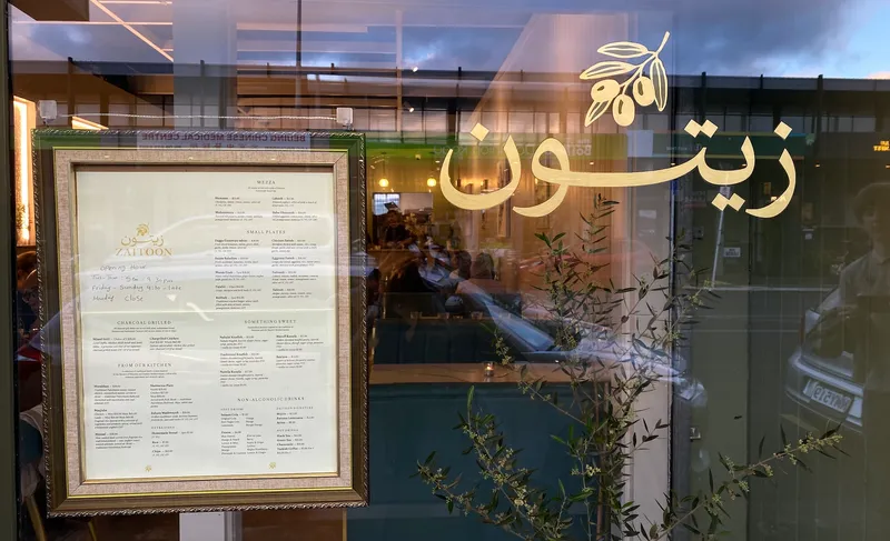 The glass frontage of a restaurant with Arabic script and an olive leaf motif in gold, next to a gold-framed menu.