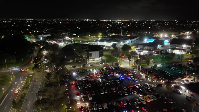 An aerial nighttime photograph showing a sprawling, crowded parking lot filled with illuminated cars and gathered people at Māngere Town Centre during a Toa Samoa fan rally.
