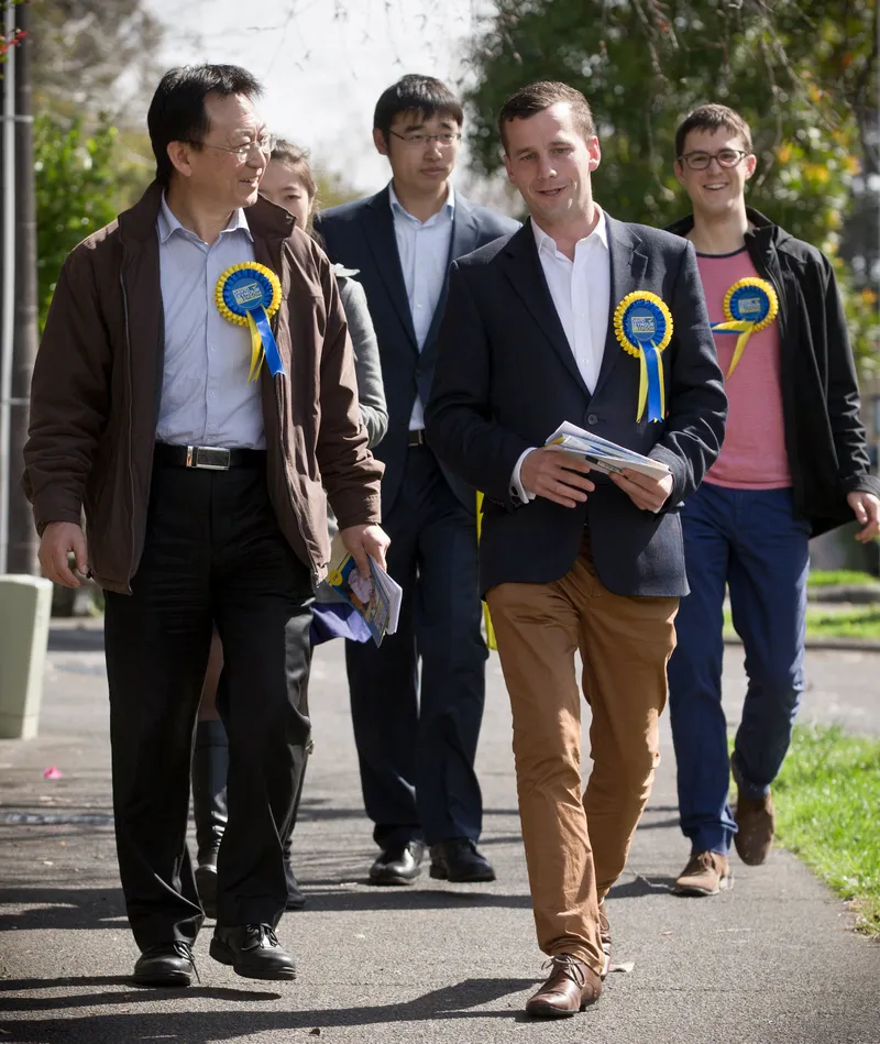 A group of people wearing rosettes and holding pamphlets walk down a suburban footpath.