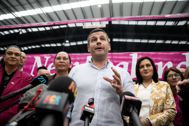 A low angle shot of a man talking in front of microphones. In the background are two women and a large pink bus.