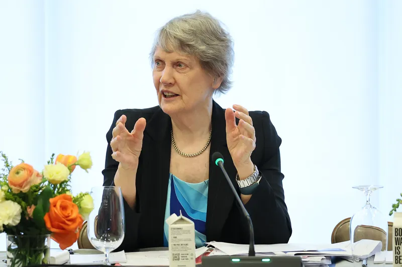 Helen Clark, a politician with grey hair and a black suit, talks while seated at a table with her hands in the air.