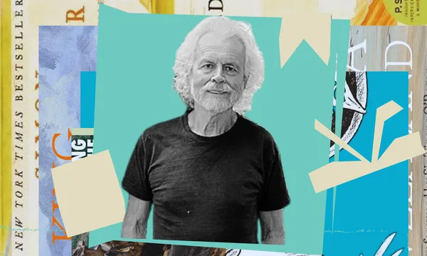 A black and white picture of Richard Wolfe who is an older man with white hair. He's wearing a black t-shirt. Behind him is a collage of coloured shapes.