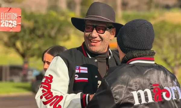 Te Pāti Māori co-leader Rawiri Waititi smiles while greeting another person wearing a black Māori-branded jacket. Waititi wears a wide-brimmed black hat, sunglasses and a black-and-white varsity jacket.