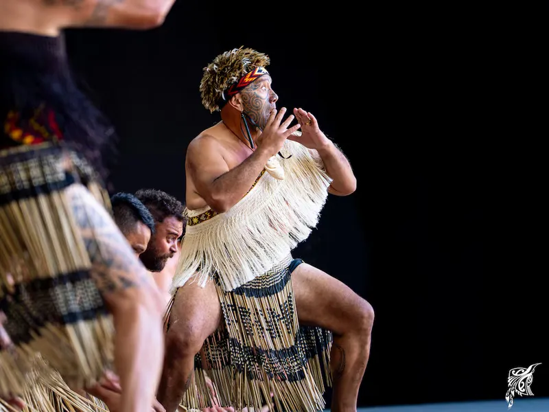 Te Pāti Māori co-leader Rawiri Waititi performs a haka with Te Taumata o Apanui at Te Matatini, wearing traditional kapa haka attire, a feathered headpiece and a piupiu, with one knee raised and both hands held near his face.