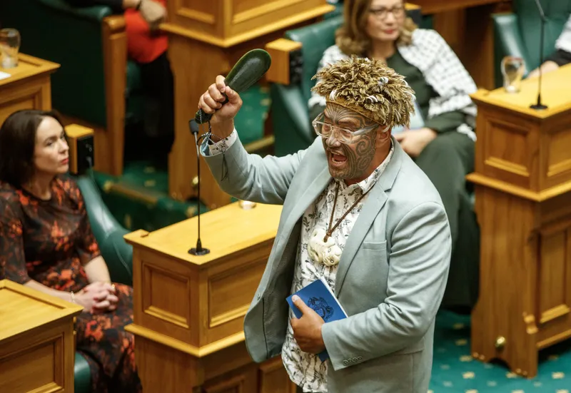 Te Pāti Māori co-leader Rawiri Waititi raises a greenstone mere and calls out in Parliament, wearing a feathered headpiece, light grey suit and glasses, with his mataora prominently visible.