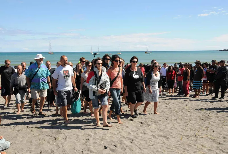 Skippers, crew and activists from a five-boat flotilla walk barefoot across the sand at Whangaparāoa Bay, with a crowd gathered behind them and yachts anchored offshore.