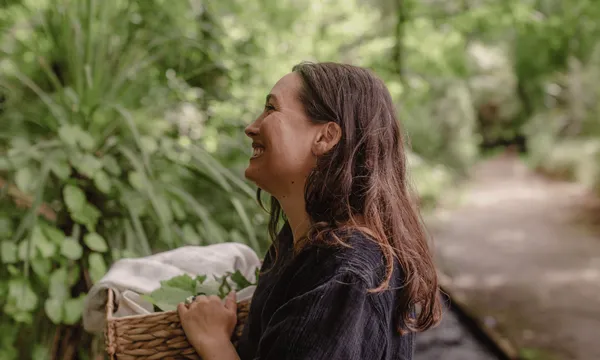 a young woman with long, straight brown hair holds a woven basket filled with green leaves and smiles off camera. she is outside in a garden setting